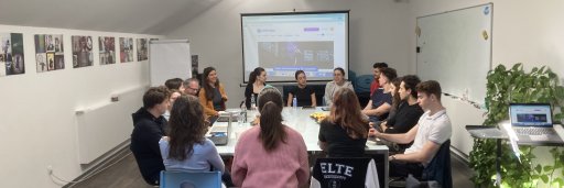 A large group of people sitting around a long meeting table during a presentation. A projection screen at the front displays the "Centropa" website in a room with slanted ceilings and skylights.
