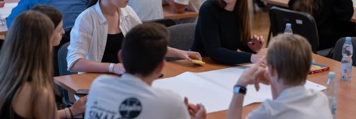 **Alt Text:**  A close-up view of a group of students seated around a wooden table in a workshop setting, actively collaborating around a large, blank sheet of paper. Other participants in the background can be seen working at nearby tables.