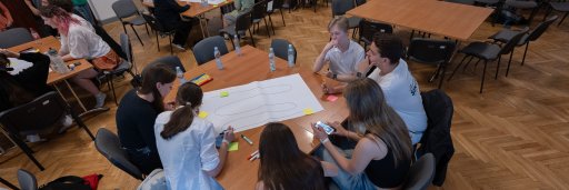 An elevated overview shows several groups of students sitting at wooden tables in a large event hall, working collaboratively on projects. The young people are discussing and writing on large sheets of paper. In the foreground, a group sits focused around a paper with sketches visible on it.