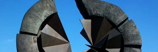 Photo of the Staro sajmište Memorial, a large abstract bronze memorial sculpture composed of two curved, fragmented forms facing each other, with angular geometric shapes at their centers, set against a bright blue sky on the bank of the Sava River in Belgrade.