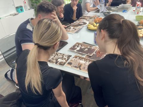 A group of young adults seated around a conference table leaning over and closely examining old black-and-white family photographs. Fruits and drinks are spread across the table.