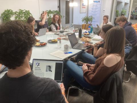 A high-angle shot of a group working around a large conference table with open laptops, papers, and snacks. In the foreground, a person works on a laptop displaying a profile template with the placeholder text "Vardas Pavardė."