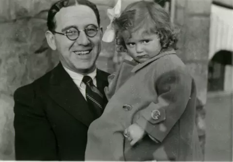 Black-and-white photograph of a smiling man wearing round glasses and a suit, holding a young girl in a buttoned coat with a bow in her hair.