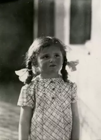 Black-and-white photograph of young Judit Kinszki with braided hair tied with large bows, wearing a patterned dress and standing outdoors beside a building.