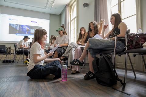Young school Students sitting in a seminar room, one girl is sitting on the floor in front of three other girls, talking with each other