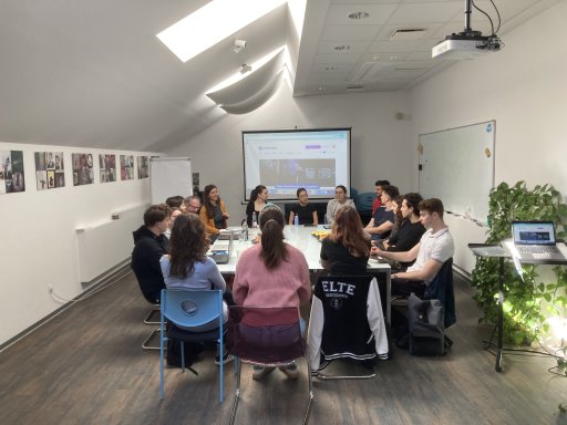 A large group of people sitting around a long meeting table during a presentation. A projection screen at the front displays the "Centropa" website in a room with slanted ceilings and skylights.