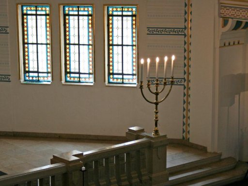 A tall, golden seven-branched menorah with six lit candles stands on a stone balustrade inside a synagogue or ceremonial room. In the background, light filters through three tall windows with decorative geometric stained-glass borders, illuminating the light-colored walls with blue decorative trim.