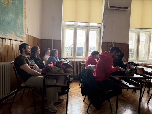 Students sitting at wooden desks in a classroom with large windows and wood-paneled walls, listening attentive to a presentation or lecture. A large map hangs on the left wall, and bright daylight fills the room through the tall windows.