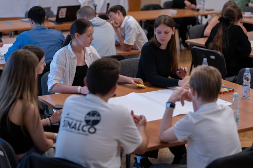 **Alt Text:**  A close-up view of a group of students seated around a wooden table in a workshop setting, actively collaborating around a large, blank sheet of paper. Other participants in the background can be seen working at nearby tables.