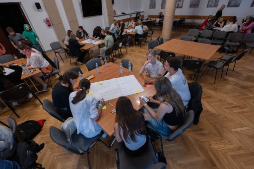 An elevated overview shows several groups of students sitting at wooden tables in a large event hall, working collaboratively on projects. The young people are discussing and writing on large sheets of paper. In the foreground, a group sits focused around a paper with sketches visible on it.